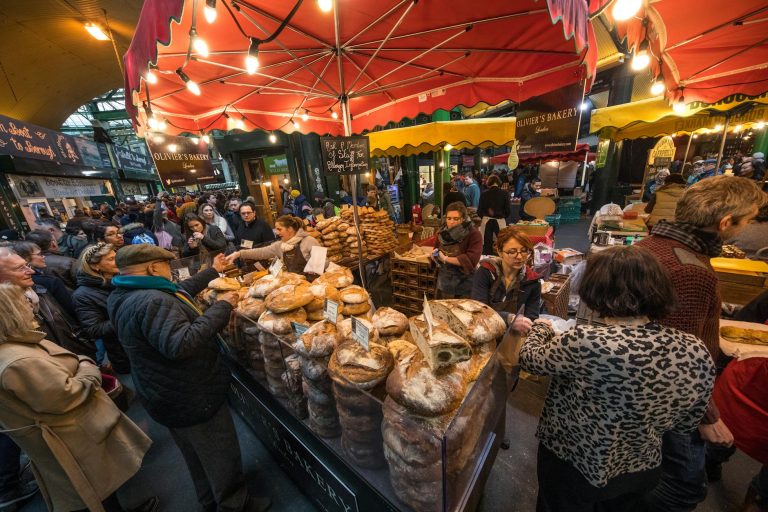 Bustling market scene at London's Borough Market with crowds buying artisan bread under colorful umbrellas.