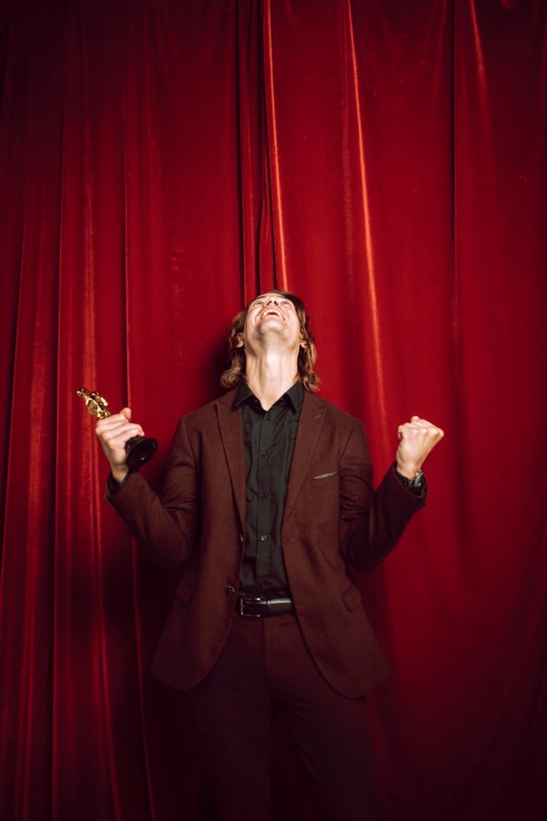 Joyful man in a suit celebrates winning an award on stage with a red curtain backdrop.