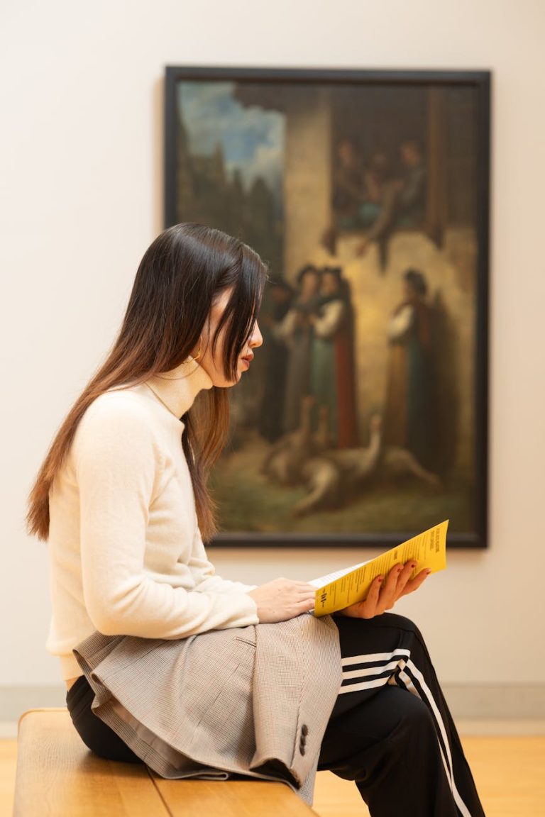 A young woman sits on a bench reading a yellow booklet in an art museum gallery.