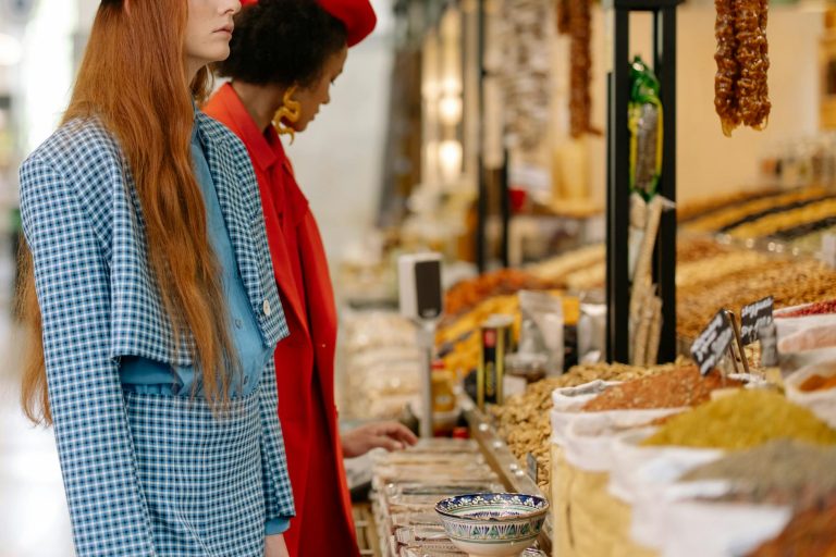 Two stylish women exploring a colorful spice market with various ingredients on display.