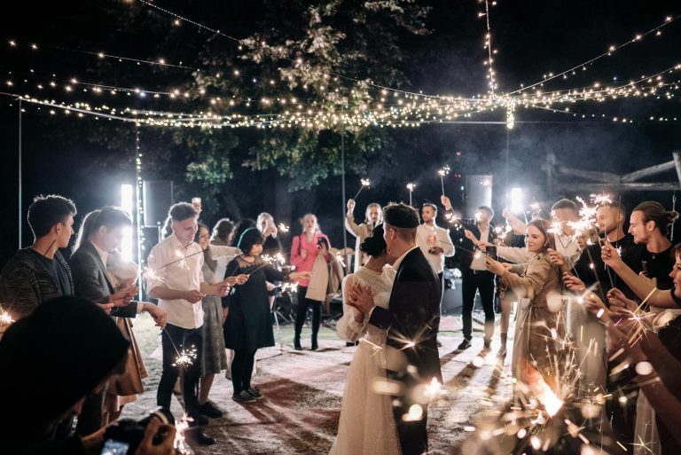 A couple dances under twinkling lights at a wedding reception, surrounded by guests holding sparklers.