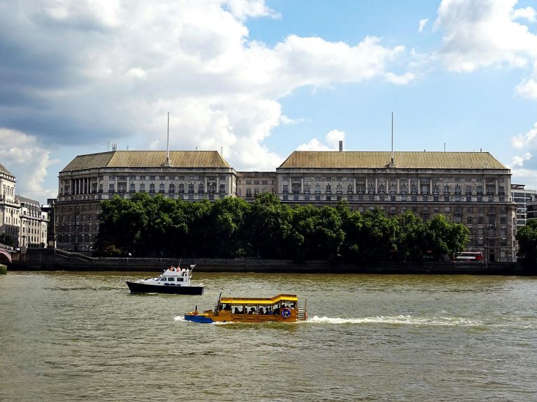 Urban scene of the Thames River in London with boats and historic architecture under a cloudy sky.