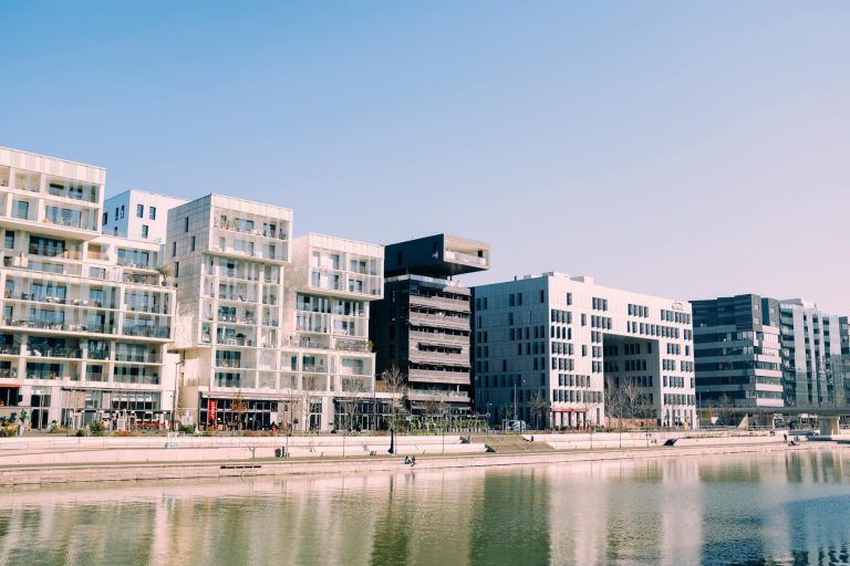 Contemporary buildings with glass facades along a waterfront in an urban setting under a clear blue sky.