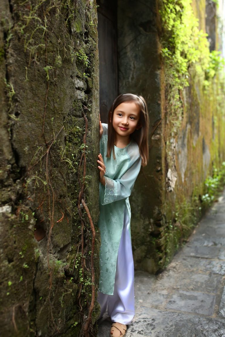 Young girl in ao dai in Hội An's mossy alleyway, peeking with a smile.