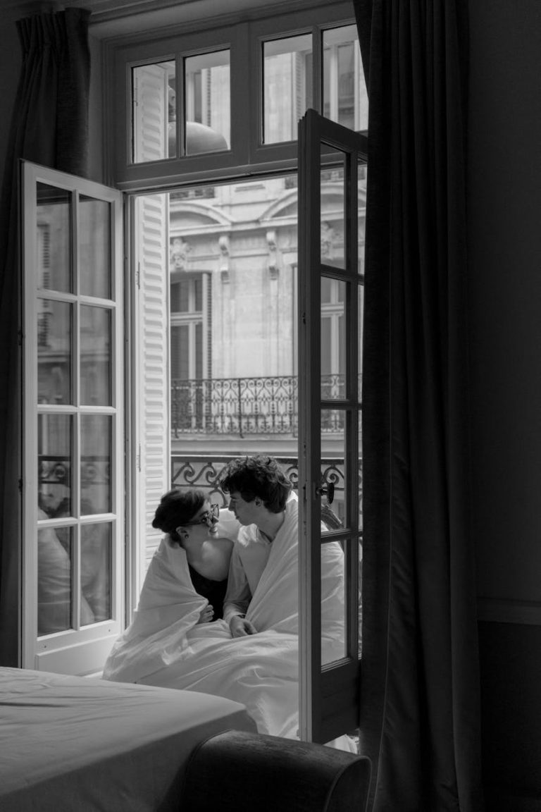 A couple wrapped in blankets sits by a window, enjoying a quiet moment together in black and white.