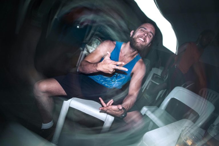 A bearded man in a tank top gestures energetically while sitting on a white chair. Blurred effect.