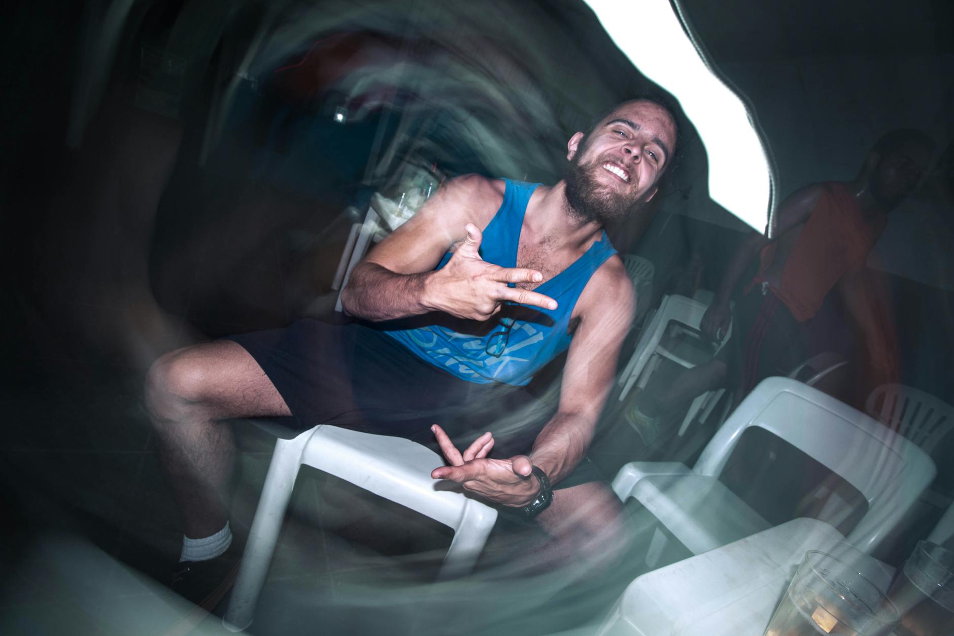 A bearded man in a tank top gestures energetically while sitting on a white chair. Blurred effect.