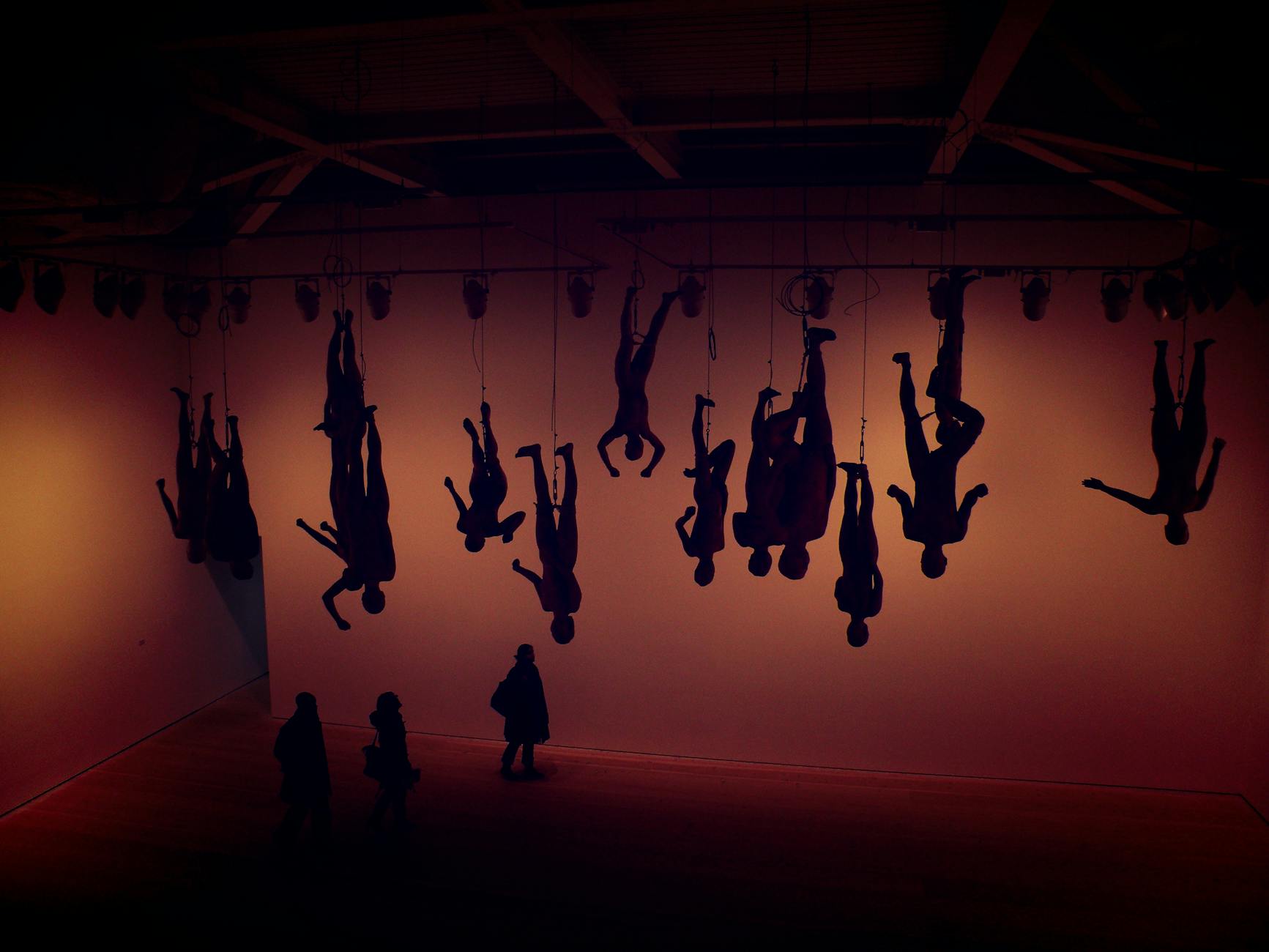 Silhouette of hanging figures in a London gallery with visitors viewing the installation.