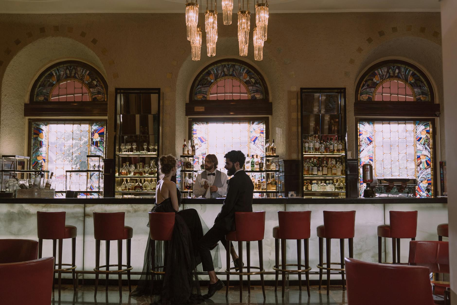 Couple in elegant attire enjoying a date at a luxurious bar with a stained glass backdrop.