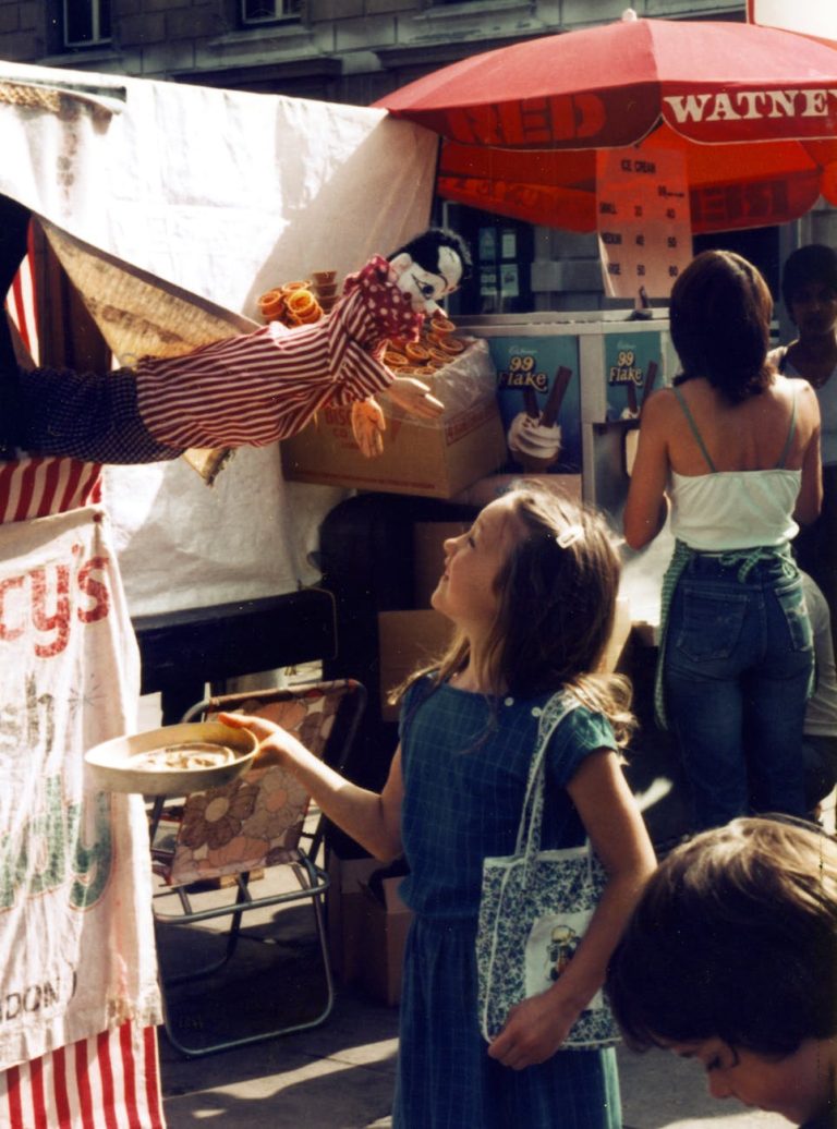 A young girl interacts with a puppet in a vibrant outdoor market setting.